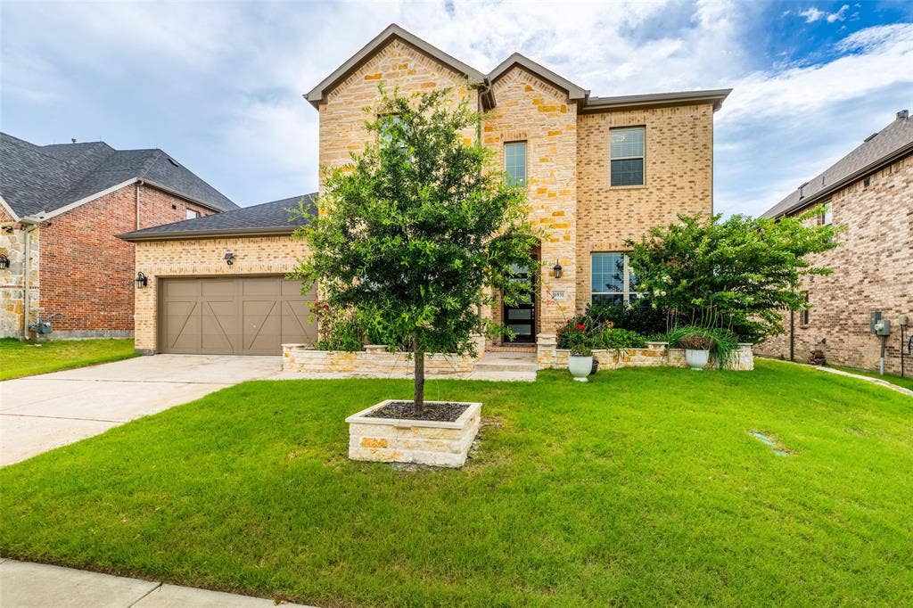 16931 Maple Bend Drive, Frisco, TX 75035 Two-story residence featuring a multi-toned brick facade, gabled roofline, integrated two-car garage with wood-finish doors, and stone-accented landscaping planters