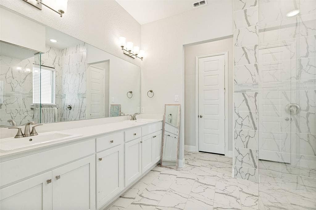 16931 Maple Bend Drive, Frisco, TX 75035 Bathroom featuring a dual-sink vanity with white cabinetry, full-width wall mirror, large-format veined tile flooring, and a walk-in shower with floor-to-ceiling tile surround