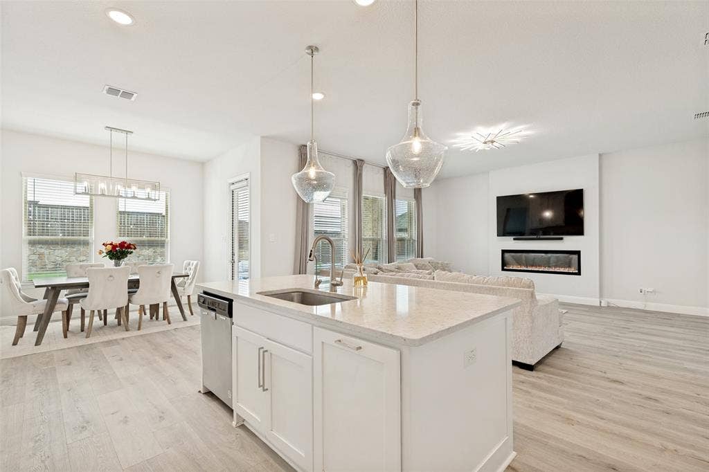16931 Maple Bend Drive, Frisco, TX 75035 Kitchen island featuring a stone countertop, under-mount sink, and integrated dishwasher, wood-finish flooring, linear electric fireplace, and multi-pendant lighting
