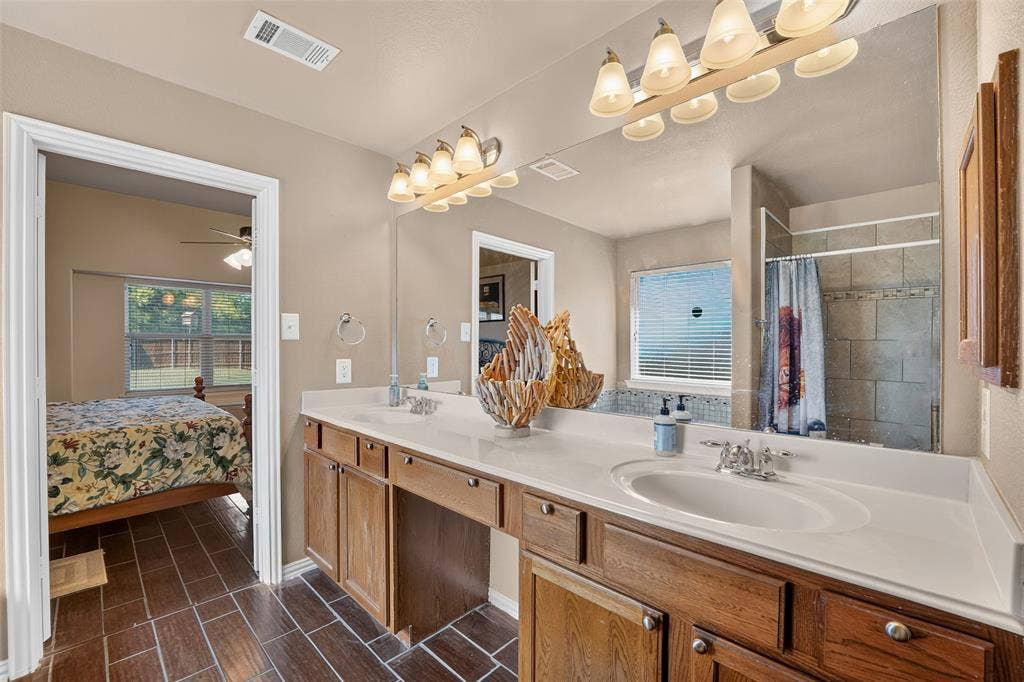 8700 Broken Bow Drive, Greenville, TX 75402 Dual-sink vanity with full-width vanity mirror, multi-light fixtures, and tiled shower and bath surround.
