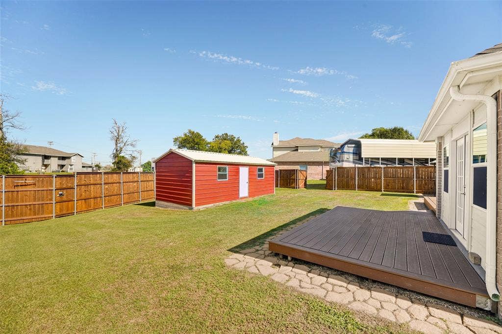 8700 Broken Bow Drive, Greenville, TX 75402 Rear exterior featuring a TREX deck, stone-style paver border, perimeter wood fencing, and a detached metal workshop. In the background you can see part of the carport and one set of double gates.
