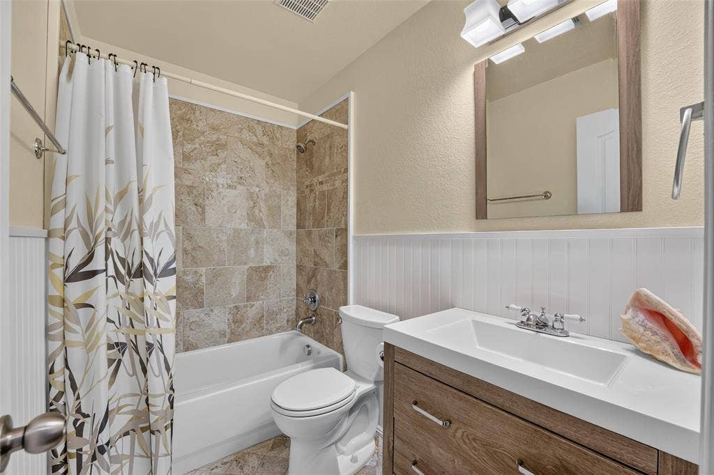 8700 Broken Bow Drive, Greenville, TX 75402 Super cute completely remodeled bathroom featuring a tub/shower combination with tile surround and flooring, wood vanity with rectangular sink, framed mirror, white beadboard wainscoting and decorative mirror and coordinated fixtures.