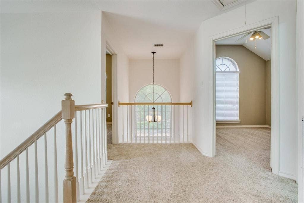 Interior landing featuring a wood-toned handrail with white balusters, carpeted flooring, a multi-light pendant fixture, and an arched window