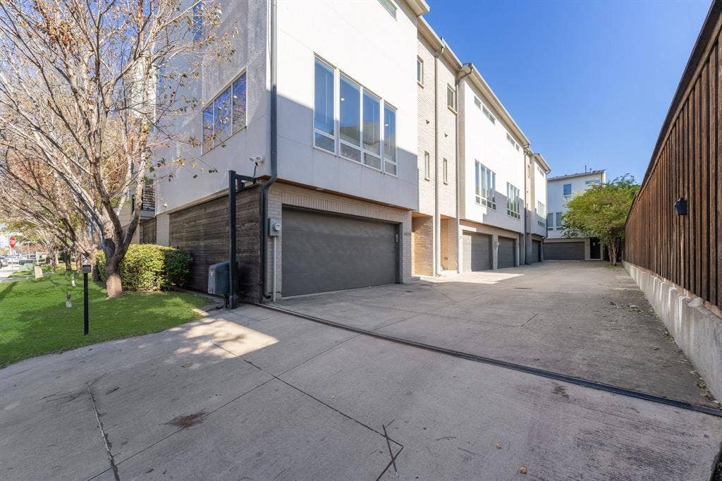 Multi-story exterior featuring a light-toned smooth facade, dark-finish metal garage doors, expansive multi-pane windows, and concrete driveway surfacing