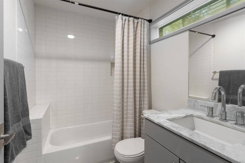 Bathroom featuring a vanity with stone countertop and integrated sink, chrome widespread faucet, horizontal clerestory window, and tub enclosure with white tile surround