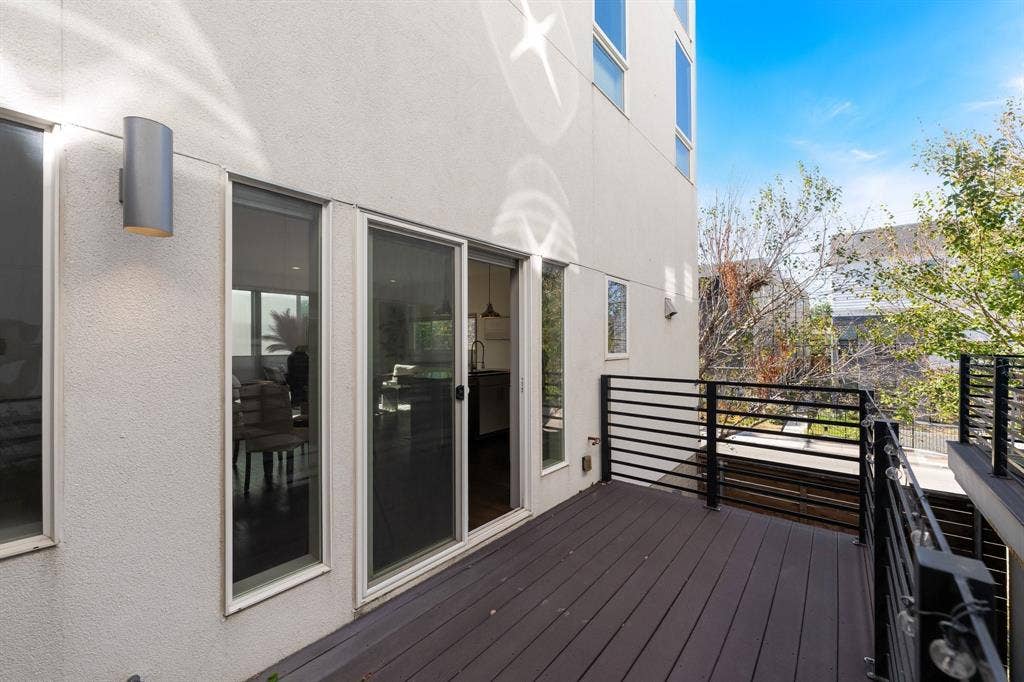 Exterior balcony featuring wood-finish decking, horizontal metal railings, sliding glass doors, and modern cylindrical wall-mounted lighting