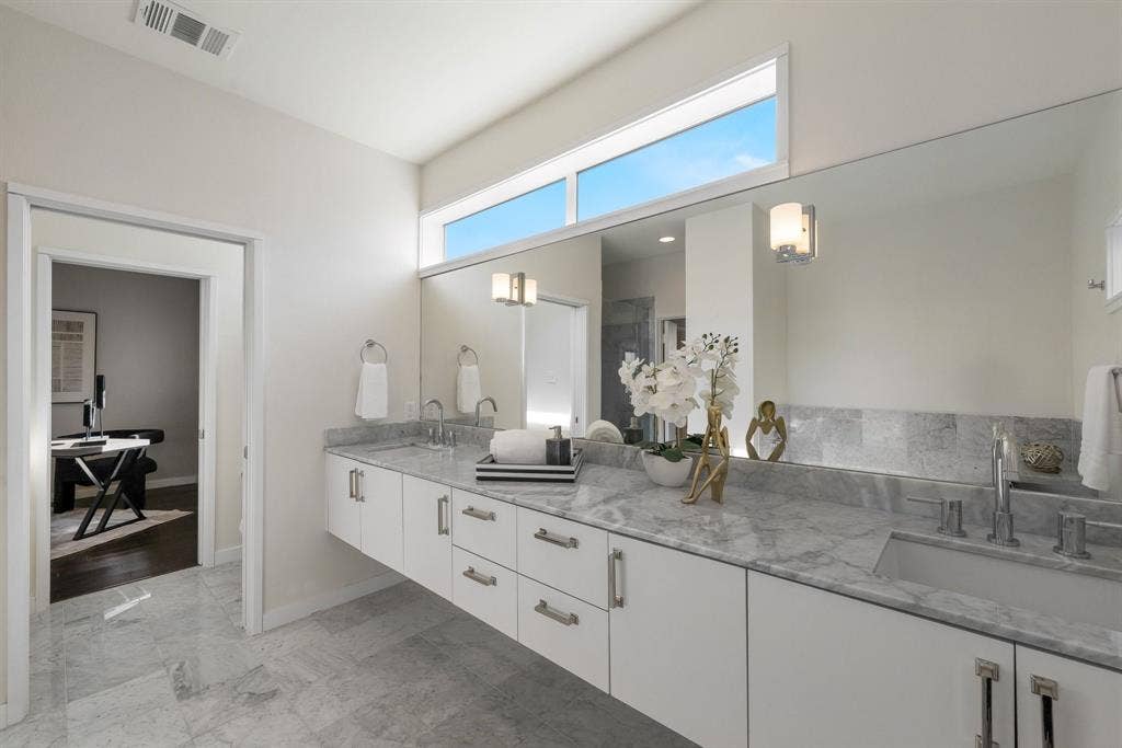 Floating vanity featuring white flat-panel cabinetry, stone countertops, dual rectangular undermount sinks, and a full-width mirror with dual-light wall sconces, complemented by high-set transom windows and stone tile flooring