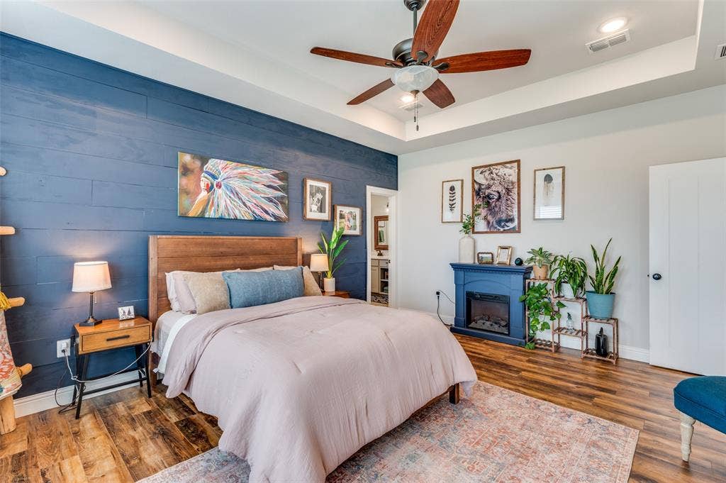 Wood-finish flooring, blue horizontal plank accent wall, tray ceiling with a multi-blade ceiling fan, and a built-in electric fireplace with a decorative mantel
