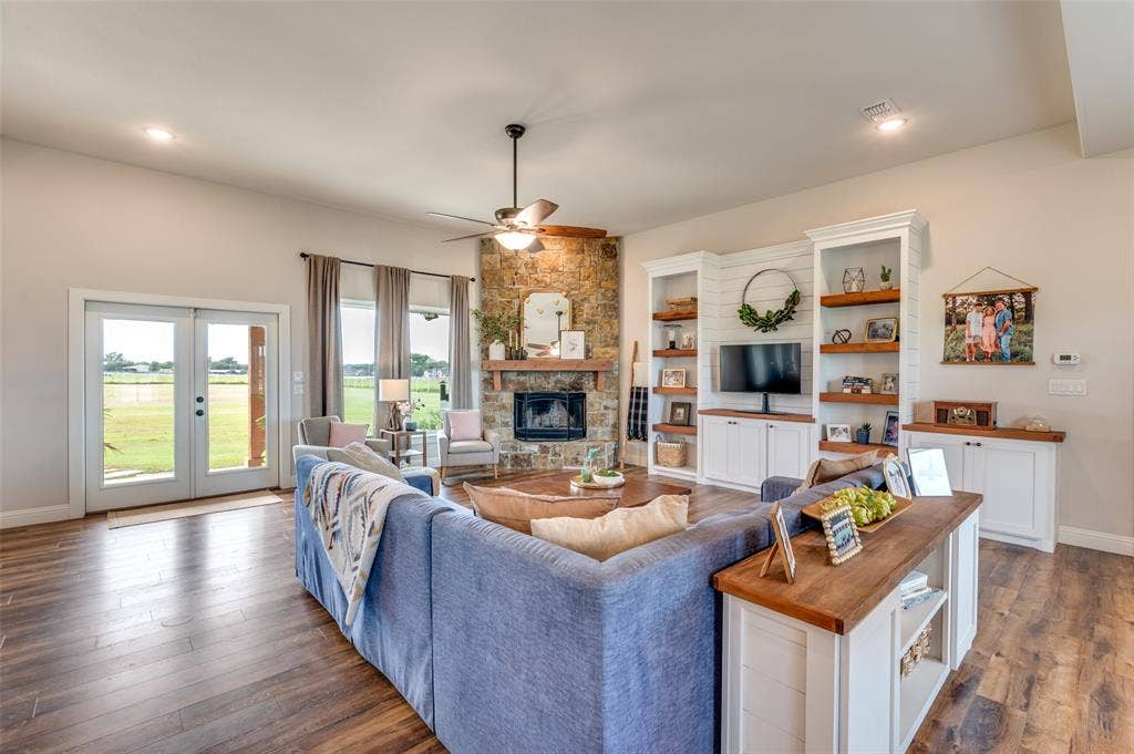 Living area featuring stone-clad corner fireplace with timber mantel, built-in cabinetry with shelving, wood-finish flooring, ceiling fan, and double glass-panel doors