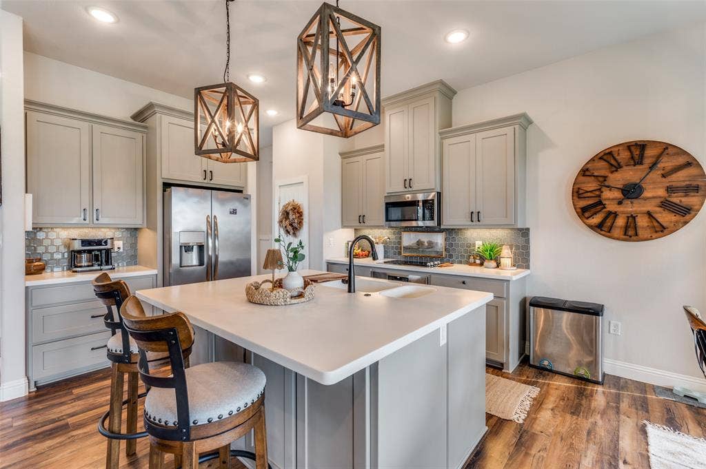 Kitchen featuring light grey shaker-style cabinetry, a central island with a solid surface countertop, stainless steel appliances, geometric pendant lighting, and hexagon-patterned backsplash tile