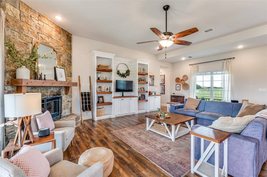 Living area featuring a floor-to-ceiling natural stone fireplace with a wood mantel, built-in white cabinetry with open shelving, wood-finish flooring, and a ceiling fan with integrated lighting