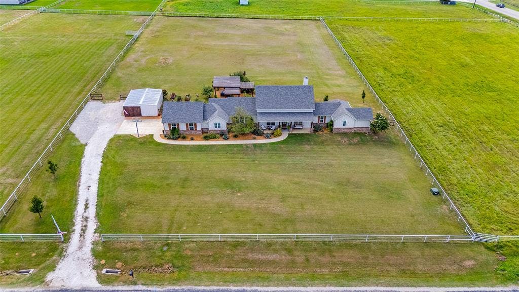 Aerial perspective of a sprawling property featuring a multi-gabled residence, a detached metal-sided structure, an expansive fenced lawn, and a graveled driveway
