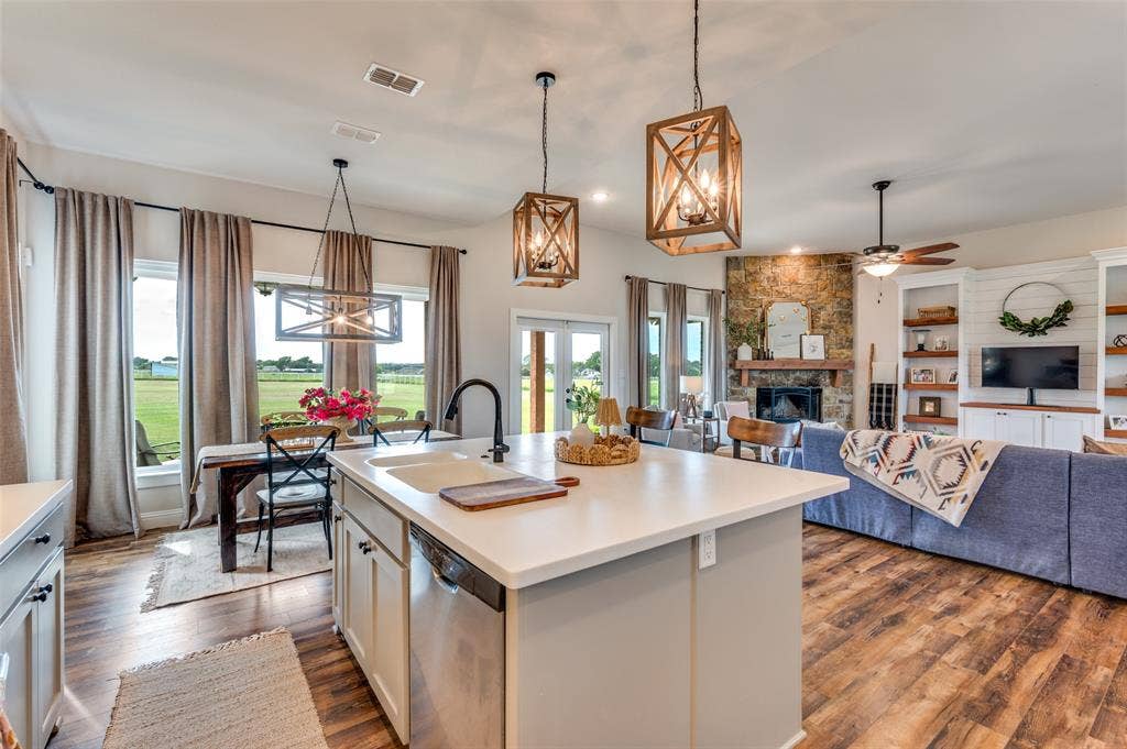 Kitchen island featuring a solid-surface countertop and a matte black faucet, open-concept layout with wood-finish flooring, stone-veneer fireplace with a wood mantel, and geometric lantern-style pendant lighting