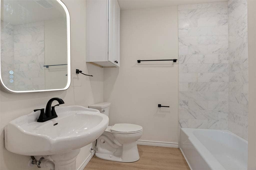 417 Keats Circle, Irving, TX 75061 Bathroom featuring a pedestal sink with matte black faucet, wall-mounted backlit mirror, built-in cabinetry, and a tub with stone-finish tile surround
