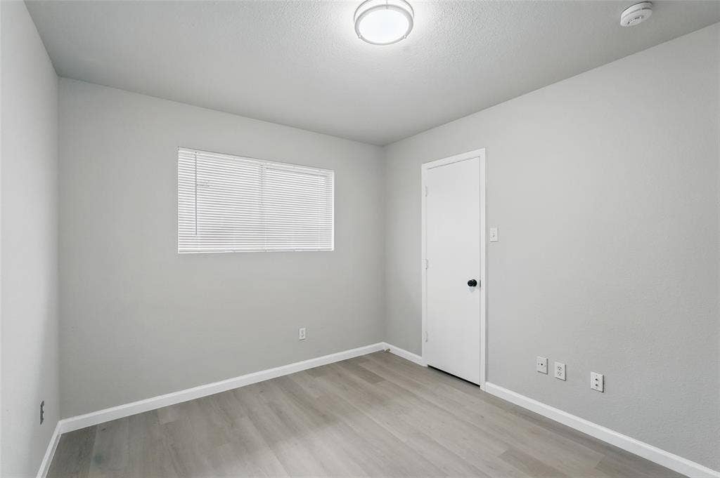 Neutral-toned room featuring wood-finish flooring, a single-panel interior door with a matte black handle, a horizontal window with adjustable blinds, and a flush-mount dome light fixture