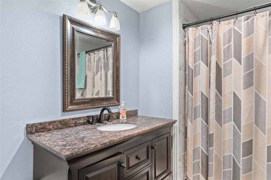 1219 Green Tree Acres Road, Kemp, TX 75143 Dark wood vanity featuring a stone countertop, an integrated oval sink, a wall-mounted framed mirror, a three-light fixture, and matte black hardware