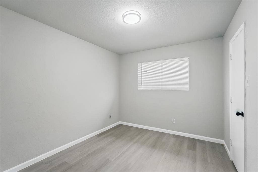 Neutral interior space featuring wood-finish flooring, light gray walls, a flush-mount ceiling light, a horizontal window with blinds, and a paneled interior door with matte black hardware