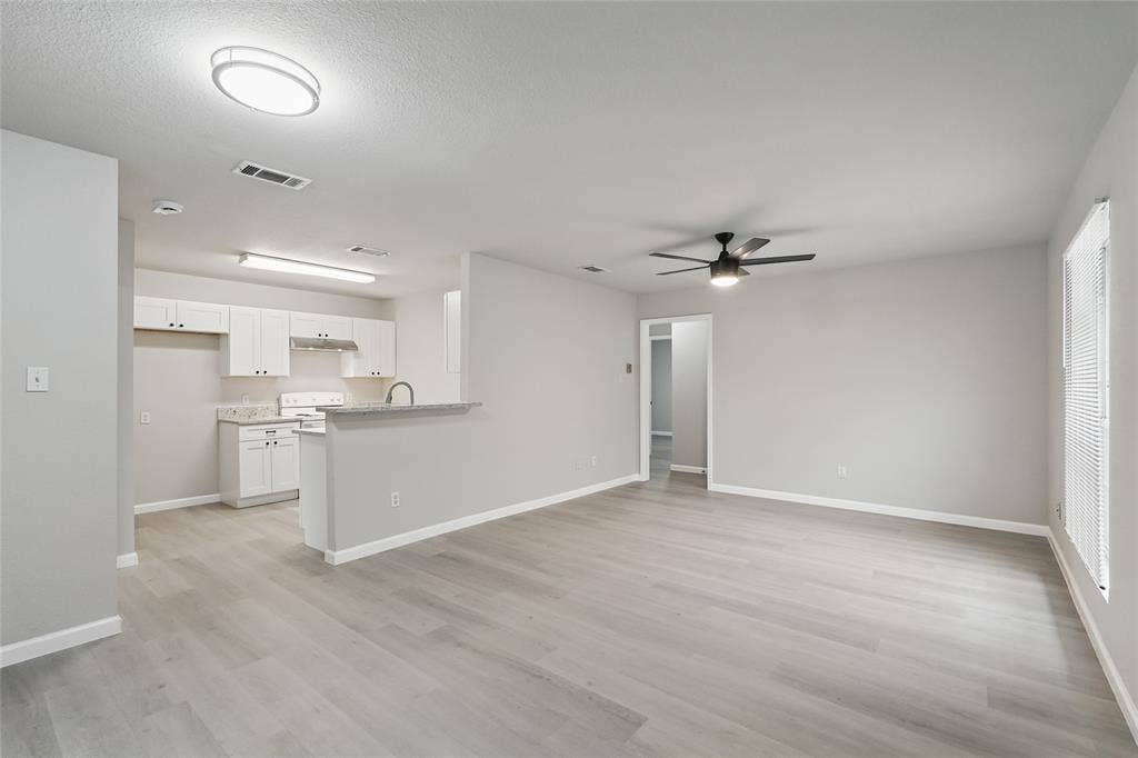 Open-concept living space featuring wood-finish flooring, white shaker-style cabinetry, a stone-surface breakfast bar, a ceiling fan with integrated lighting, and flush-mount overhead illumination