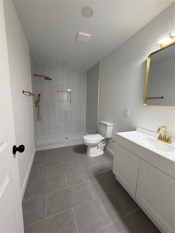 Bathroom featuring a walk-in shower with vertical tile surround and built-in recessed niche, white vanity cabinet with a integrated sink, gold-tone plumbing fixtures, and large-format neutral floor tile