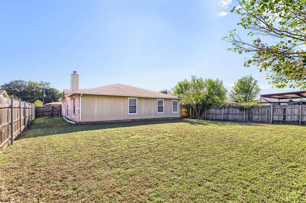 2039 Angelica Way, Dallas, TX 75217 Expansive backyard featuring a grass lawn, full-perimeter wood fencing, a masonry chimney, and a metal-roofed carport structure
