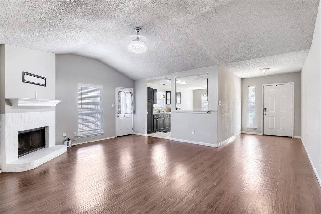 2039 Angelica Way, Dallas, TX 75217 Vaulted ceiling featuring a fireplace with a white brick surround and mantel, wood-finish flooring, and natural light provided by multiple windows and glass-paneled exterior doors