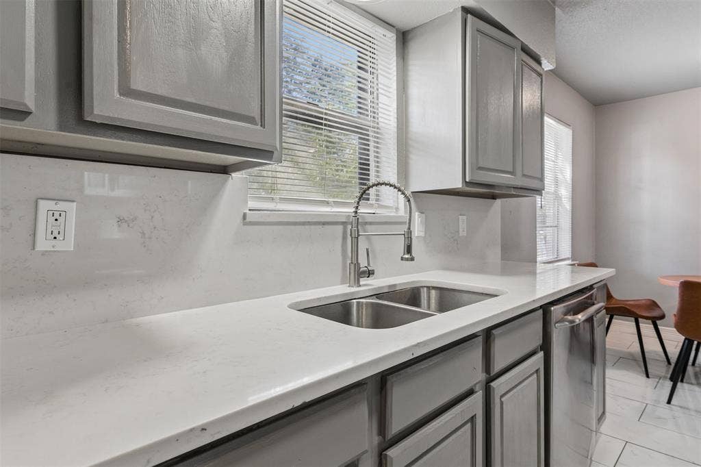 Kitchen featuring grey shaker-style cabinetry, white countertops, a full-height stone-finish backsplash, an undermount stainless steel double-basin sink with a gooseneck faucet, and stainless steel dishwasher