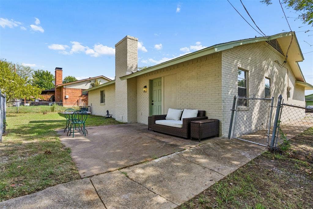 Exterior featuring light-toned brick masonry, a concrete patio, an external chimney, and chain-link fencing