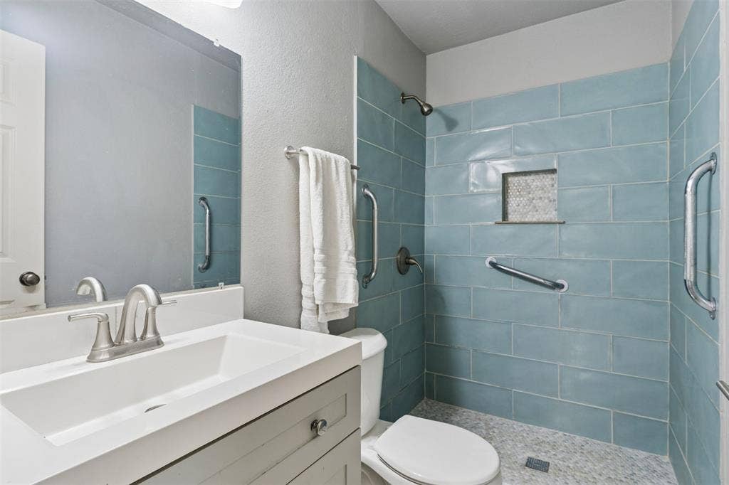 Bathroom featuring a white vanity with a rectangular sink, large wall-mounted mirror, walk-in shower with blue subway-style tile, built-in recessed niche, and stone-finish flooring