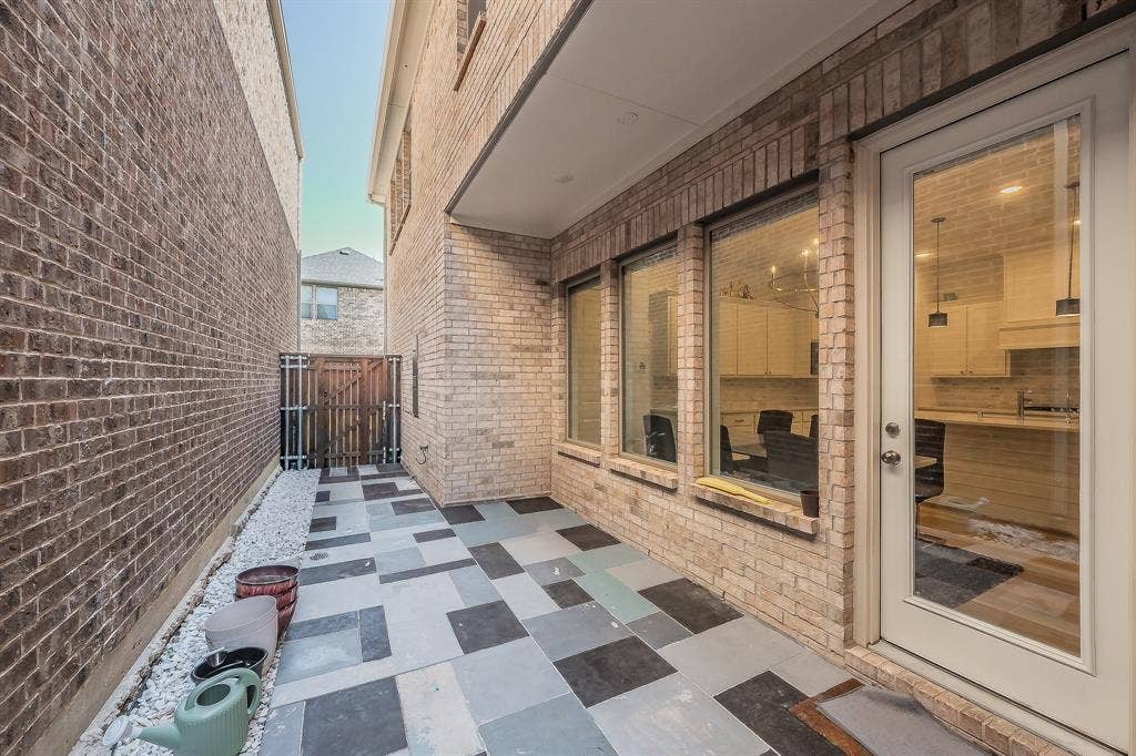 Exterior side patio featuring multi-toned stone tile pavers, brick masonry walls, a glass-panel entry door, and a pebble-stone garden border