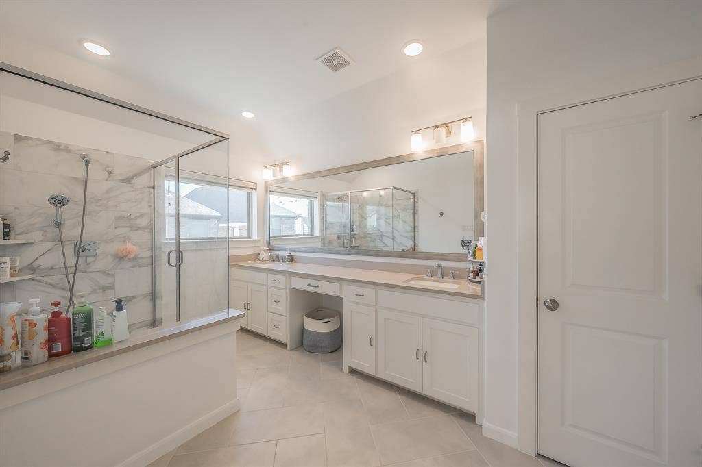 Dual-sink vanity with light-finish cabinetry, stone-surface countertops, full-width wall mirror, glass-enclosed shower with stone-tile surround, and recessed ceiling lighting and private toilet in the primary bathroom