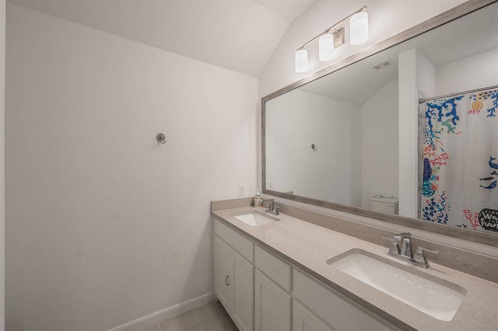 Double vanity secondary full bathroom featuring a light-toned solid surface countertop, dual undermount sinks, white Shaker-style cabinetry, a large framed vanity mirror, and three-light wall-mounted fixture