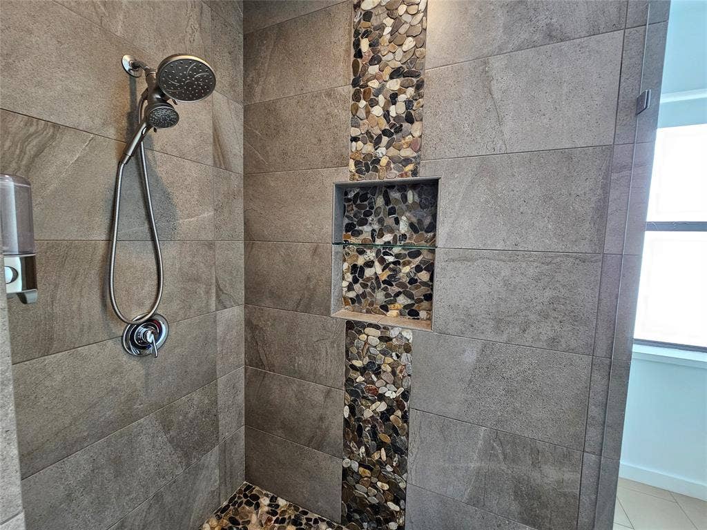 4215 Roseland Avenue, Dallas, TX 75204 Walk-in shower featuring large-format ceramic tile, a vertical decorative pebble-stone inlay, built-in recessed shelving, a brushed-metal handheld shower fixture, and a natural stone floor