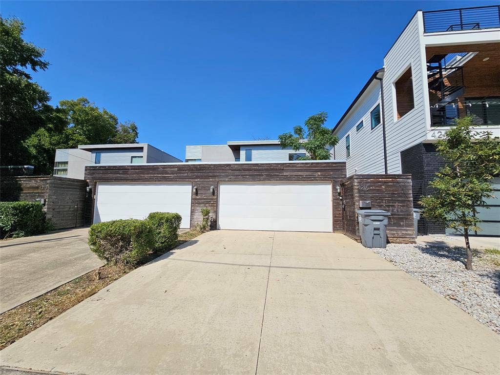 4215 Roseland Avenue, Dallas, TX 75204 Modern exterior facade featuring horizontal wood siding, two white garage doors, concrete driveway, and a third-level balcony with metal railing