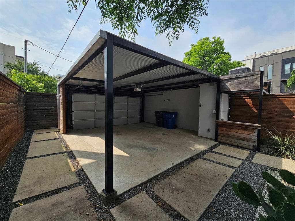 4215 Roseland Avenue, Dallas, TX 75204 Covered parking structure featuring a steel frame, corrugated metal roof, concrete slab flooring, and integrated wood-accented service area, enclosed by horizontal slat wood fencing and stone-paver hardscaping