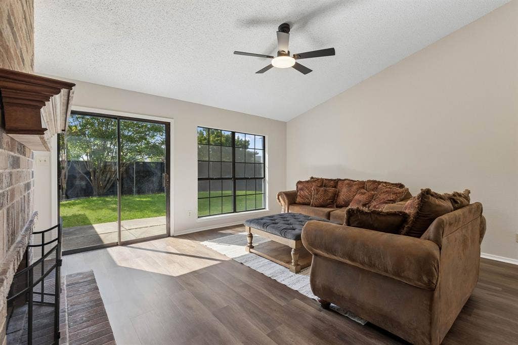 Vaulted ceiling living area featuring a floor-to-ceiling brick fireplace, wood-finish flooring, a multi-blade ceiling fan with integrated lighting, and grid-pane windows
