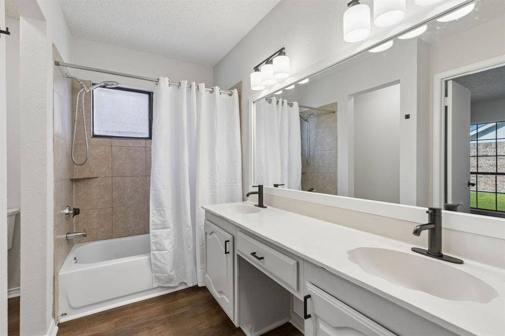 Double vanity featuring a white countertop, dual undermount sinks with matte black faucets, wood-finish flooring, and a full-width wall mirror with integrated lighting