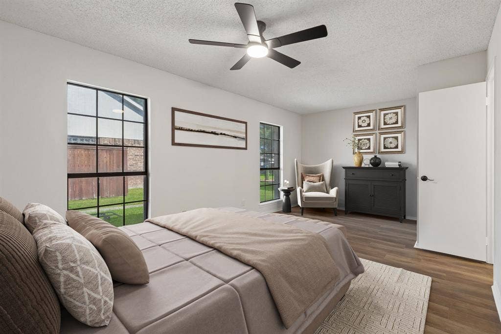 Room featuring wood-finish flooring, dual windows with black frames, and a modern multi-blade ceiling fan with integrated lighting.  Furniture is virtually staged.