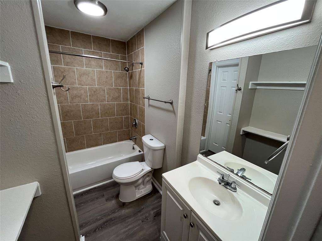Bathroom featuring wood-finish flooring, vanity with integrated sink, rectangular mirror with integrated lighting, and tile-surround shower and tub combination