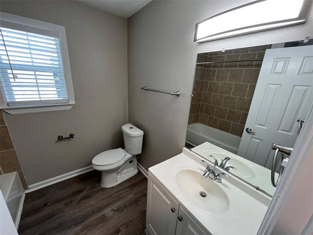 Bathroom featuring a white vanity cabinet, wood-finish flooring, a wall-mounted vanity mirror, a rectangular tub enclosure with tile surround, and brushed nickel hardware
