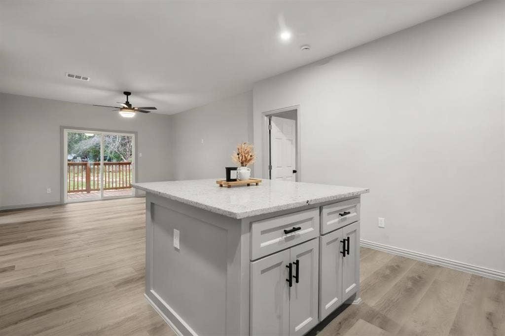 Kitchen island featuring light-toned cabinetry, light-flecked stone countertop, and matte black hardware, paired with wood-finish flooring and sliding glass doors