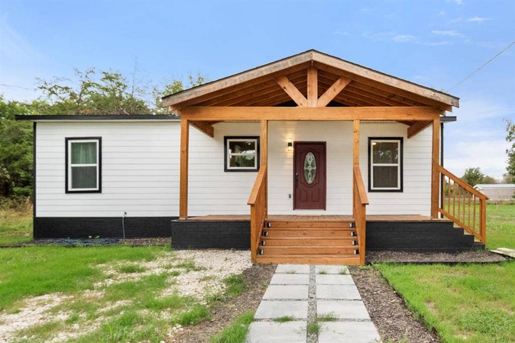 Exterior featuring white horizontal siding with a black foundation base, a covered wood-framed front porch, matching wood staircase and railing, and a stone paver walkway