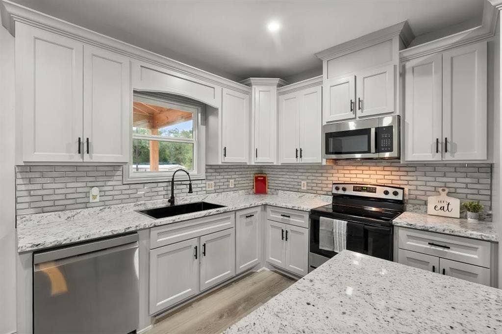 Kitchen featuring white shaker-style cabinetry, stone-finish countertops, grey subway tile backsplash, stainless steel appliances, and a matte black faucet with a coordinating deep-basin sink
