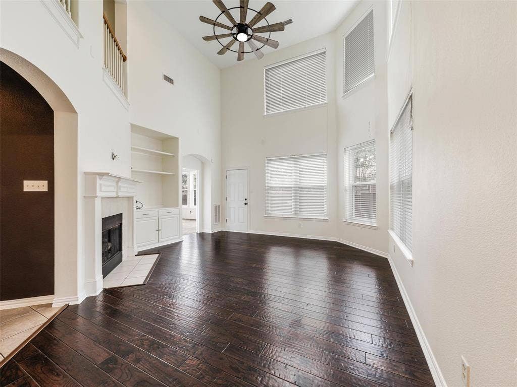 3504 Anita Drive, McKinney, TX 75070 Double-height ceiling featuring a multi-blade ceiling fan, dark wood-finish flooring, built-in cabinetry with shelving, a white fireplace mantel, and large windows