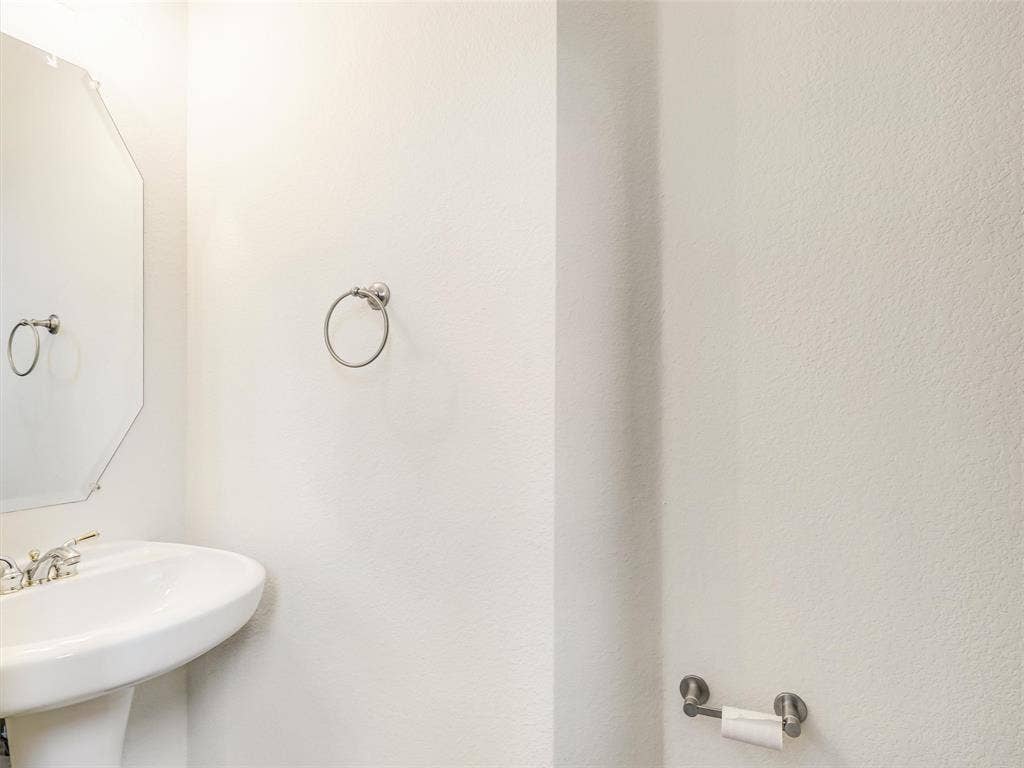 3504 Anita Drive, McKinney, TX 75070 Wall-mounted pedestal sink featuring a polished metal faucet, complemented by an octagonal wall mirror, a circular towel ring, and a coordinating toilet paper holder in a brushed metallic finish