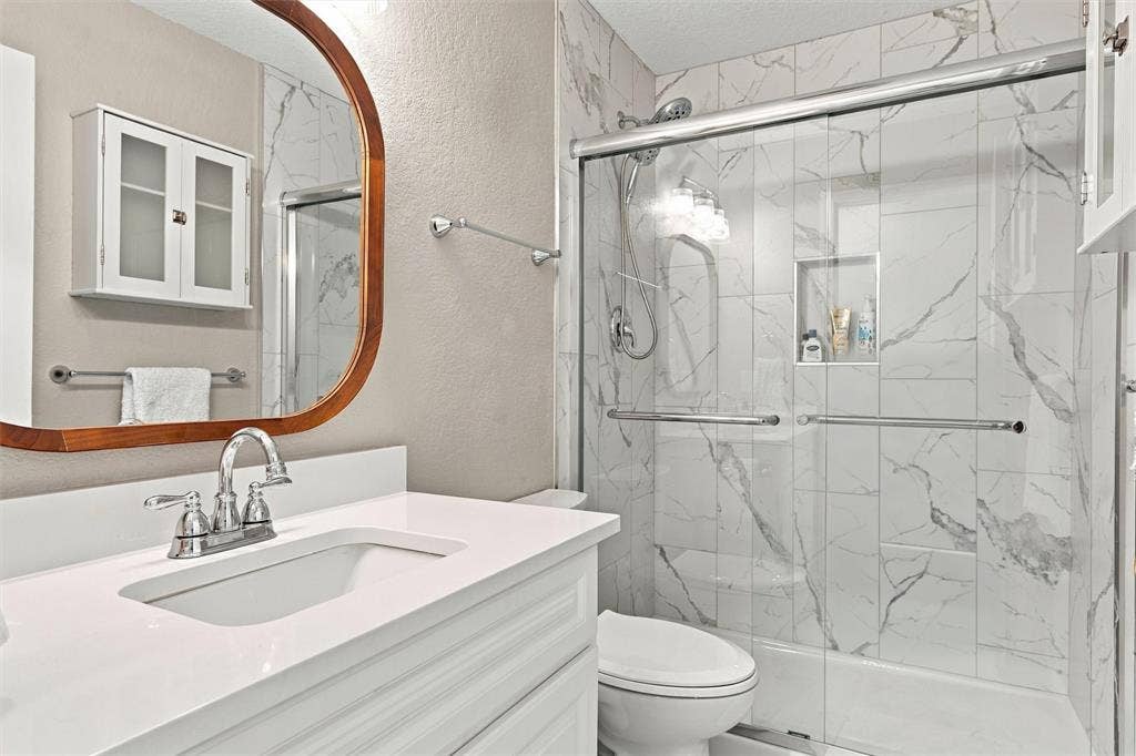 813 Chambers Creek Circle, Hewitt, TX 76643 Bathroom featuring a white vanity with a rectangular sink, quartz countertop, polished chrome faucet, arched wood-frame mirror, and shower enclosure with a sliding glass door and integrated tile niche