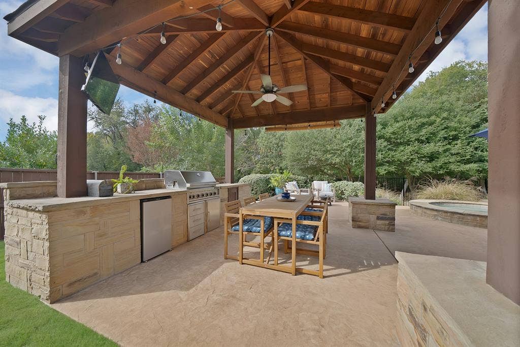 2141 Waterrock Drive, Allen, TX 75013 Covered outdoor kitchen featuring a timber-framed pavilion, stone-clad cabinetry, built-in stainless steel grill, beverage refrigerator, and concrete patio flooring