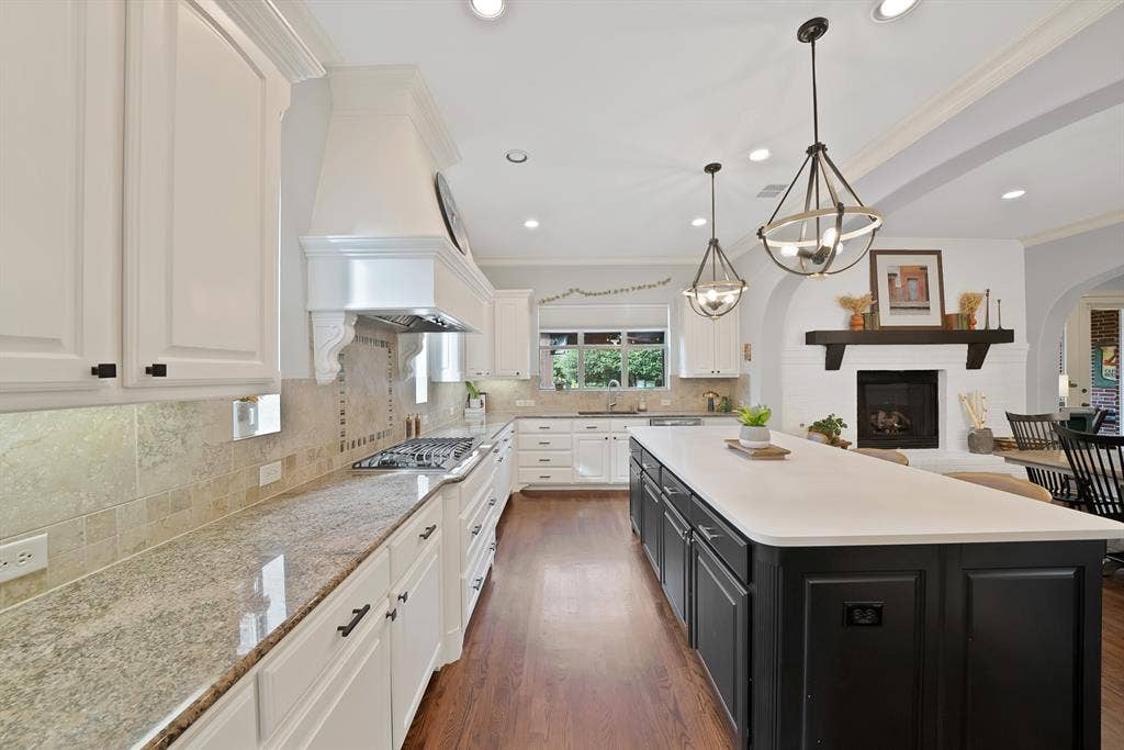 2141 Waterrock Drive, Allen, TX 75013 Kitchen featuring wood-finish flooring, a central island with a quartz countertop, stone tile backsplash, white cabinetry with a matching range hood, and a built-in fireplace with a wooden mantel