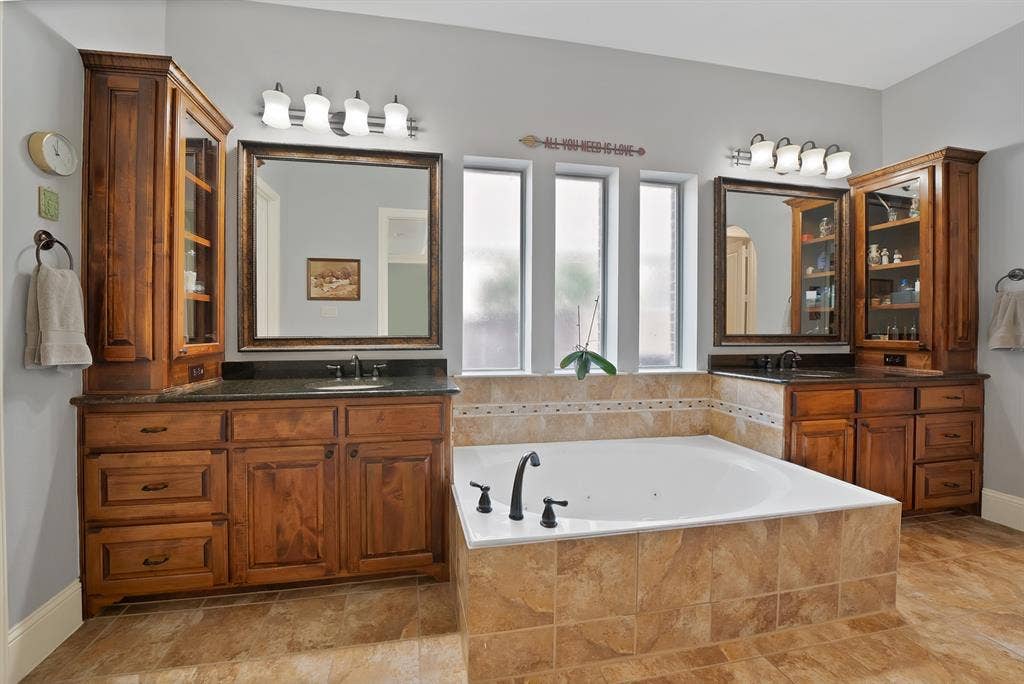 2141 Waterrock Drive, Allen, TX 75013 Dual-sink vanity with stained wood cabinetry, dark stone countertops, undermount sinks, tile-surround soaking tub, and matching wall-mounted vanity lighting