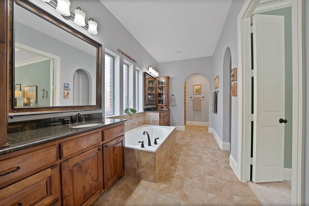 2141 Waterrock Drive, Allen, TX 75013 Bathroom featuring wood cabinetry, stone-finish countertops, a drop-in jetted tub with tile surround, ceramic tile flooring, and arched architectural entryways