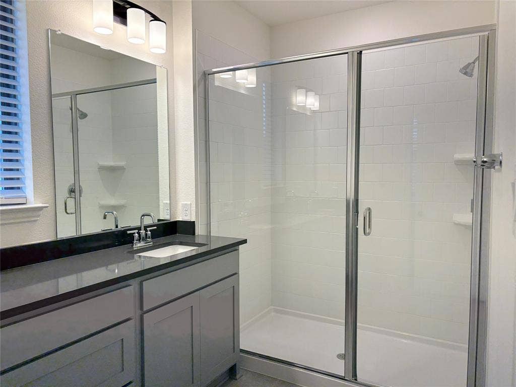 2528 Brunswick Way, Allen, TX 75013 Bathroom featuring a grey vanity with a dark countertop, a walk-in shower with white tile surround and a glass door, a large wall-mounted mirror, and a three-light fixture